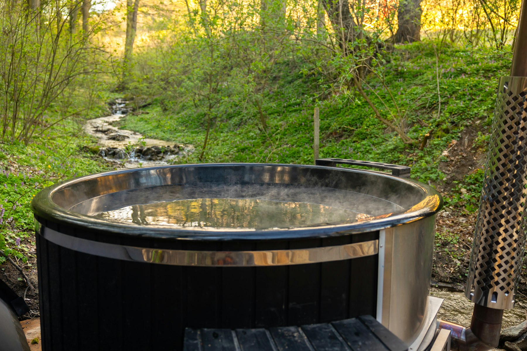 A steaming wood-fired hot tub beside a woodland stream, spring greenery and low golden light all around.