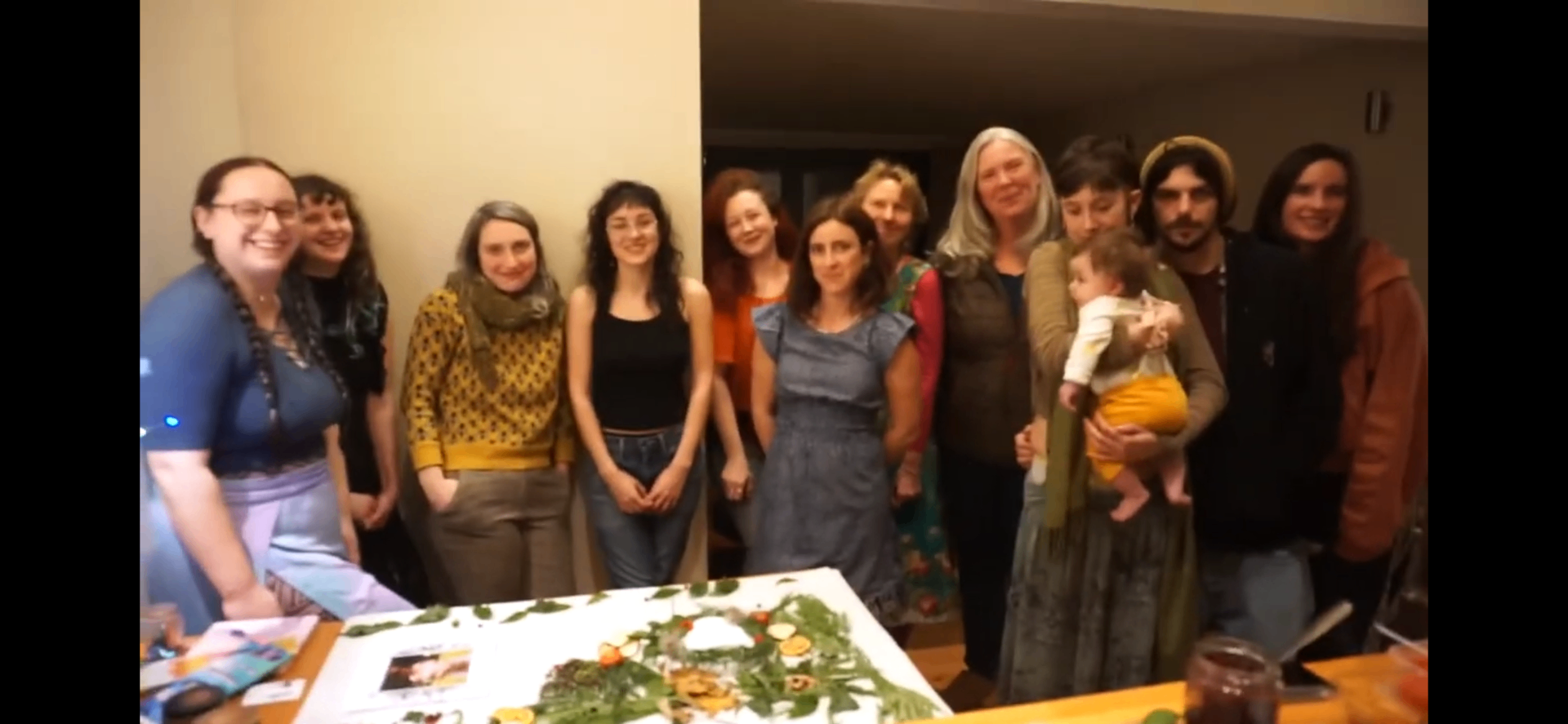 A group of sisters and a baby gathered together around a table laid with greenery and seasonal offerings.