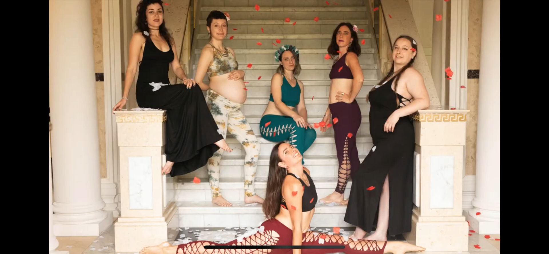 Six women on marble stairs, red petals falling around them — a celebration moment.